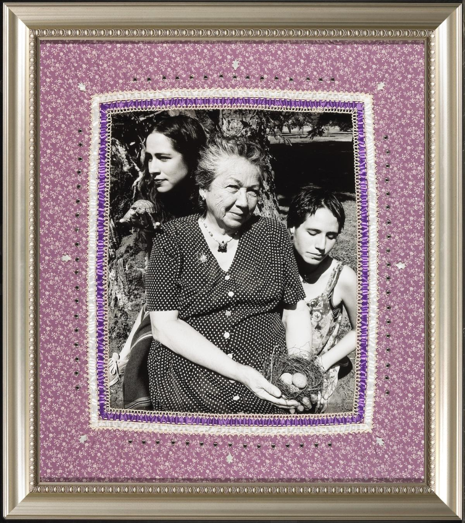 Black and white photo of an older indigenous woman in a polka dot dress holding a nest with eggs, with two younger folks behind her, surrounded by a textile border with purple and white patterning