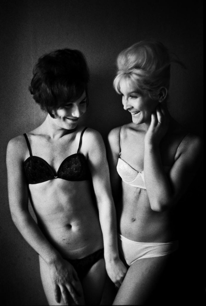 Black and white photograph of two women standing close together and smiling at one another, one with dark hair wearing a dark bra and underwear, the other with blonde hair wearing white bra and underwear