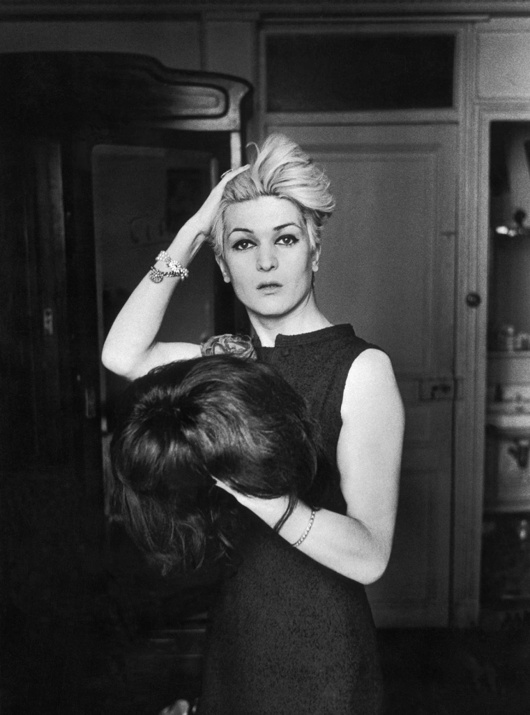 Black and white photograph of a woman running her hand through short blonde hair and looking at the camera, holding a large brunette wig in her other hand