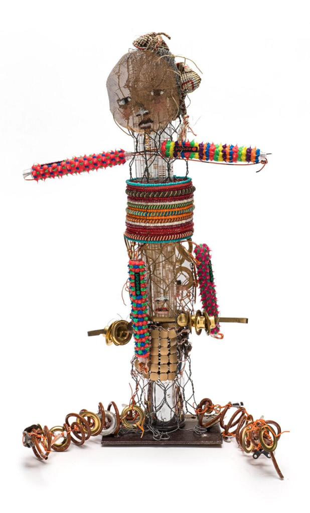 Sculptural figure made up of metal parts, colorful plastic elements, barbed wire, and striped bracelets, with a brown clay face