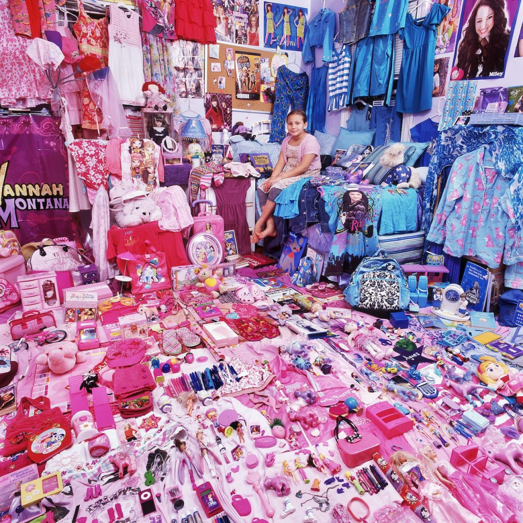 Photo of a girl sitting on her bed with all of her blue and pink possessions laid out around the floor, walls, and bed, with about 2/3's filled with pink items and 1/3 blue (mostly clothing)