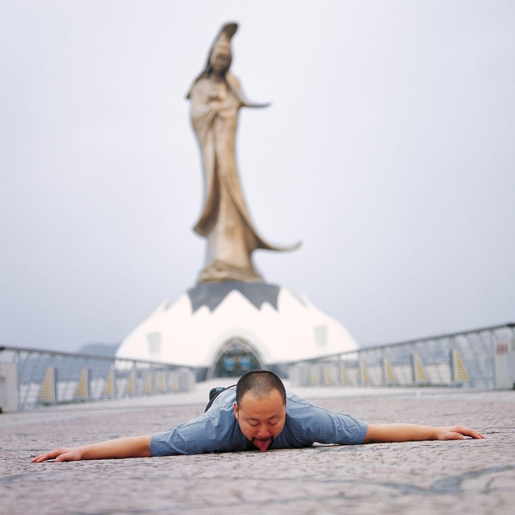 Photo of the artist in Macau lying prostrate on the ground with his tongue licking the pavement, in front of a large out of focus sculpture in the background