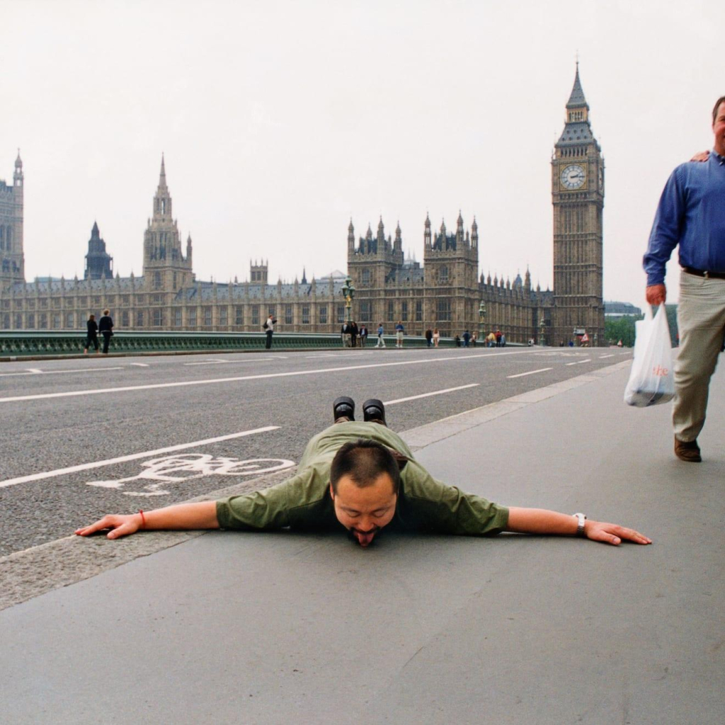 Photo of the artist in London lying prostrate on the ground with his tongue licking the pavement, in front of Big Ben and the palace