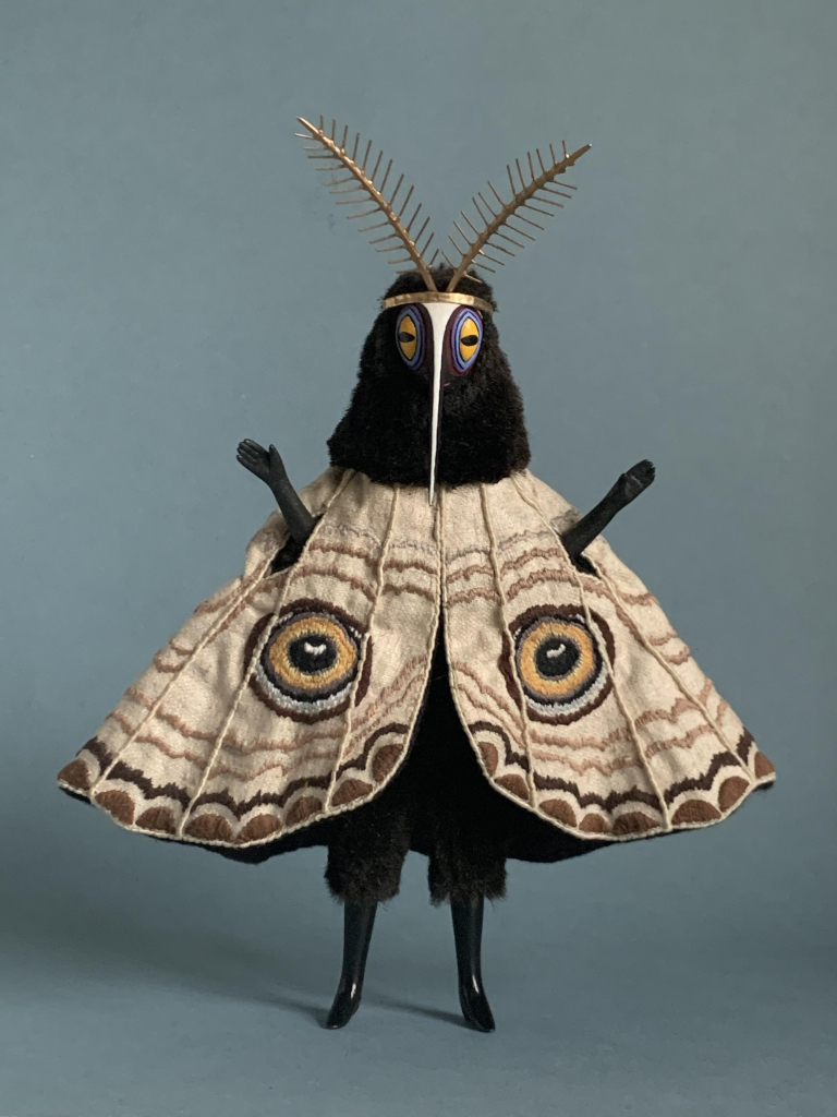 Small sculpture of a standing mothlike creature with golden antennae and a black fuzzy body, outstretched arms lifting through a cape that looks like wings decorated with eyes.