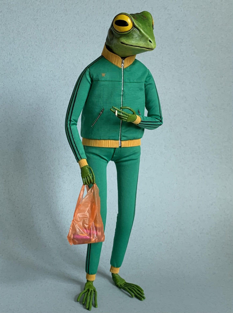 Small sculpture of a standing humanoid frog wearing a green tracksuit, holding a plastic shopping bag in one hand and a cigarette in the other