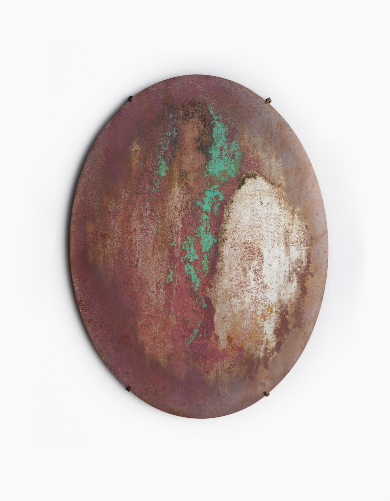 An oval metal disc hanging on a white wall like a mirror, but the surface is covered in a rust-red, cream, and green patina