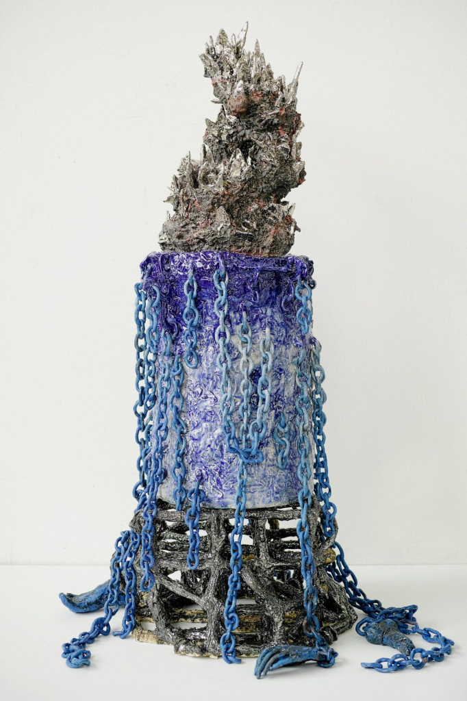 Vertical tower ceramic sculpture with long blue chains hanging down a rounded column center with blue glaze, and a spiky top section in grey and brown