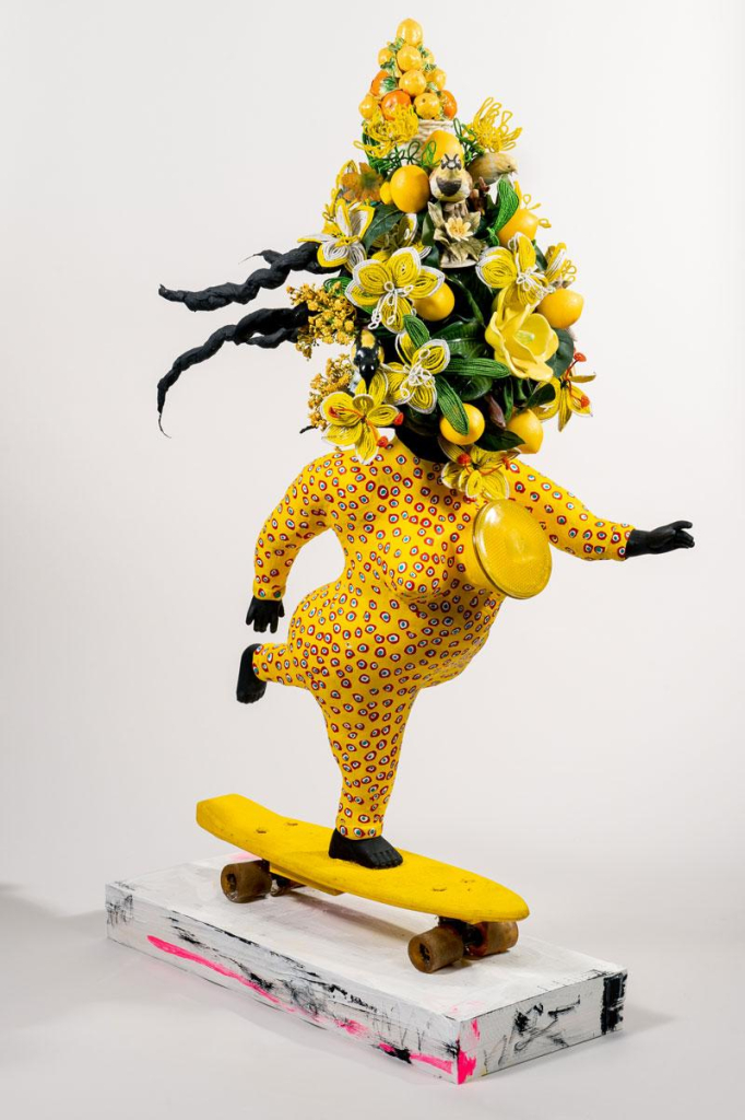 Sculpture of a bright yellow figure painted a red dot pattern, balanced on a skate board. Their head is large bouquet of yellow flowers and citrus