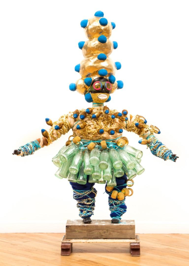 Sculpture of a female figure with a gold torso and skirt made of glass bottles. Their head is topped with a tall, bulbous gold and blue tower