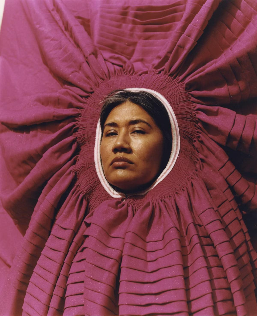 Photo of an indigenous Peruvian woman looking seriously at the camera, or face emerging through the opening of a mauve-colored garment circling around her head