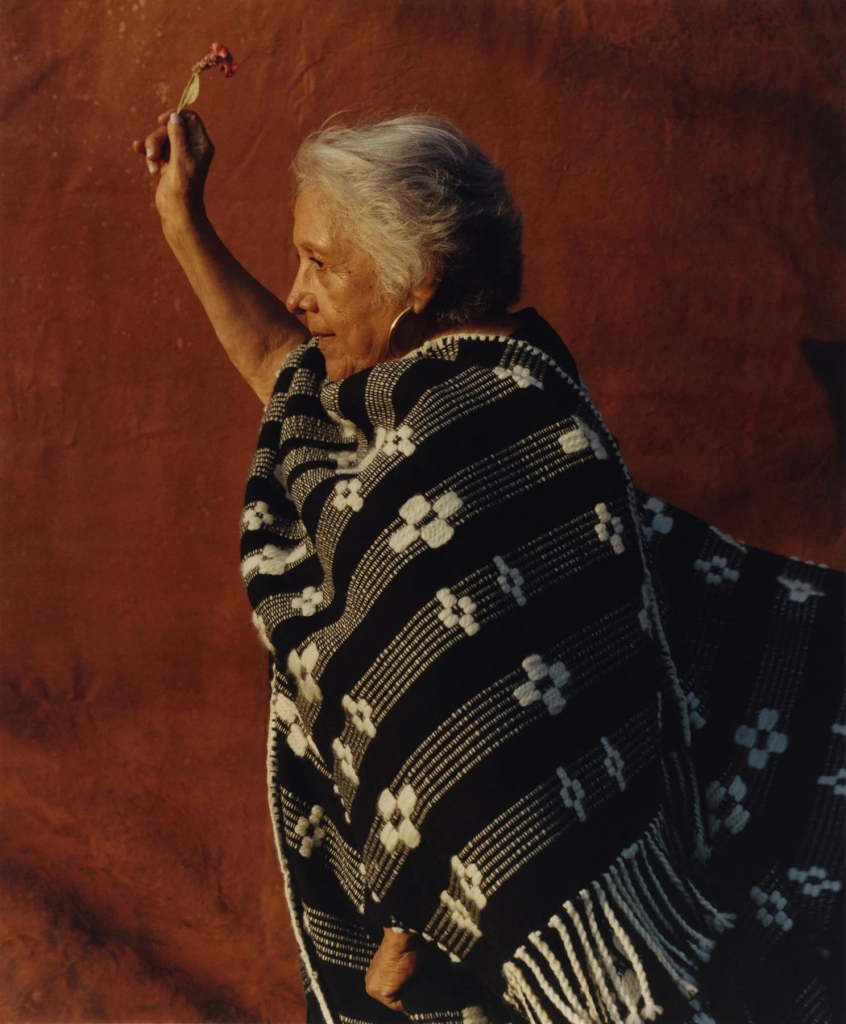 Photo of an older indigenous Peruvian woman standing in profile against a rust-colored wall, wearing a black and white blanket and holding up a small flower in her right hand