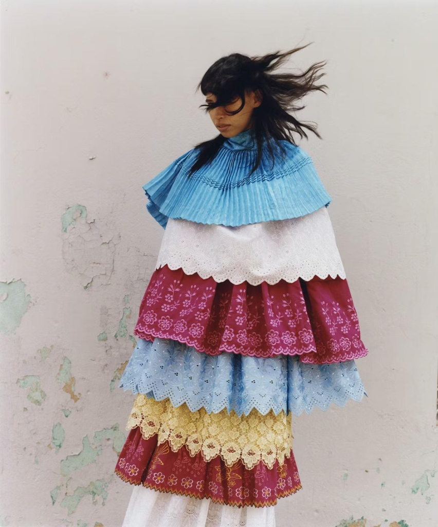 Photo of a young indigenous Peruvian woman standing against a white wall, the wind blowing her dark hair across her face. She is wearing a multi-tiered garment of blue, white, yellow, and red fabrics with scallopped edges and floral details