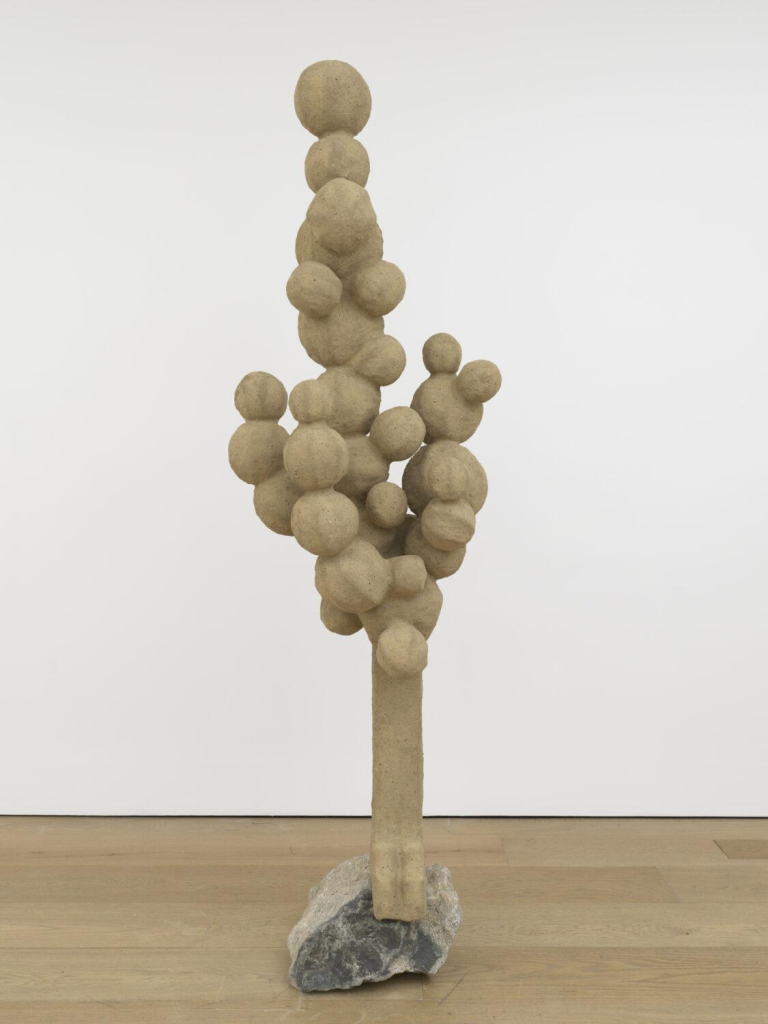 Light brown freestanding sculpture resembling a narrow tree with circular forms for leaves, attached to a rock base