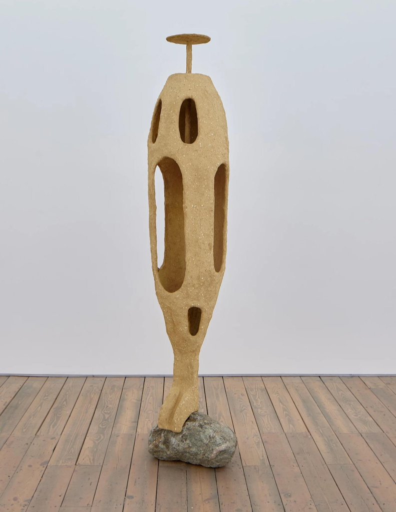 Light brown freestanding sculpture in an oval shape with narrow curved openings, attached to a rock base