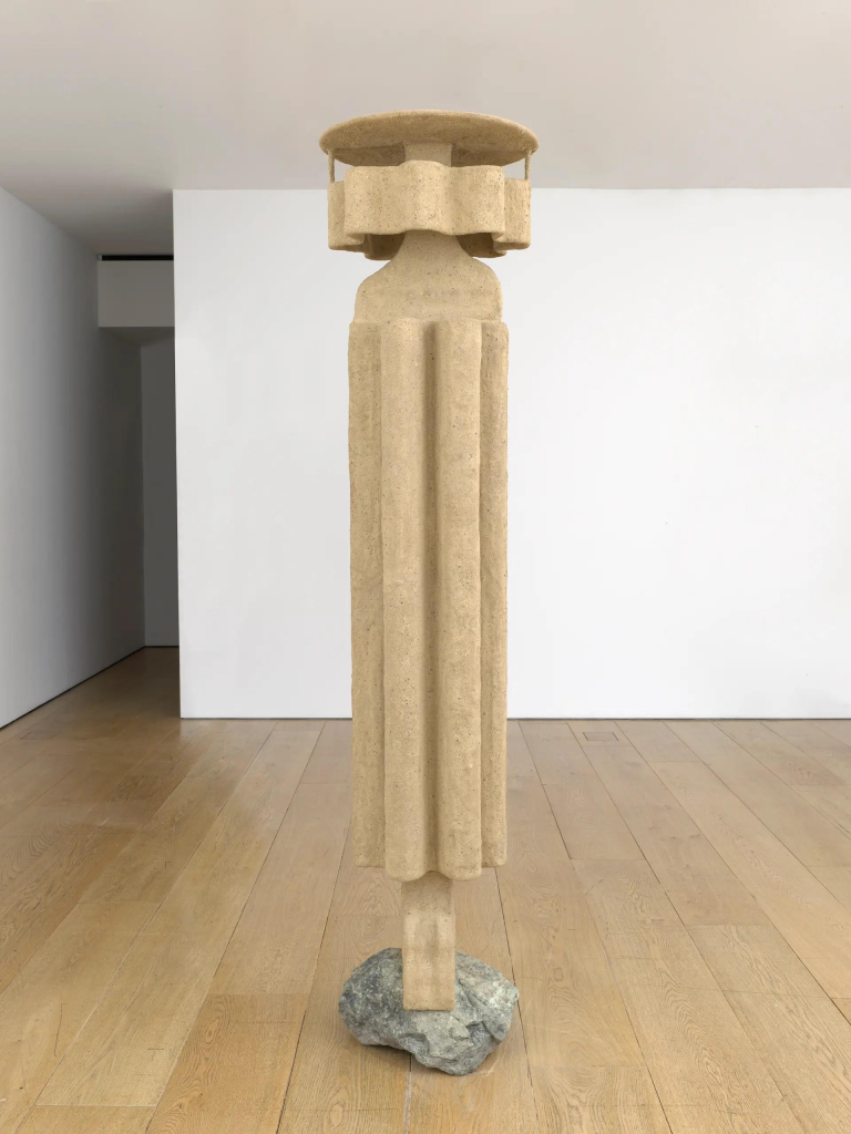 Light brown freestanding sculpture resembling a fluted column, with a thin rectangular bottom and multi-petaled circular top, attached to a rock base