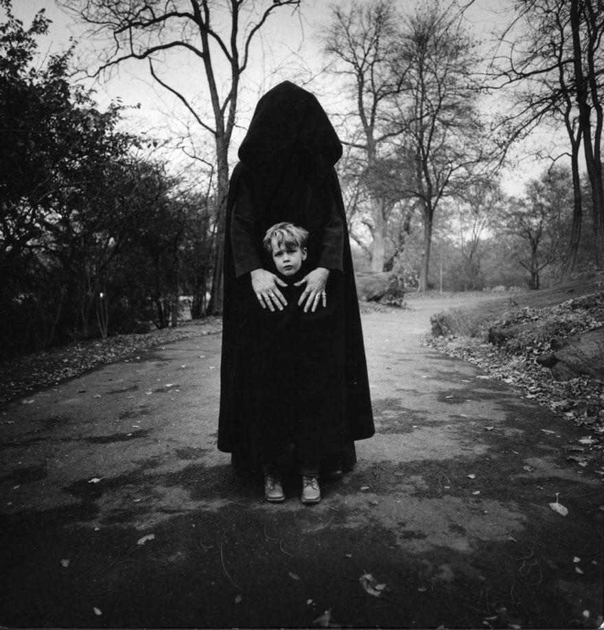 Black and white photo of a small blonde child standing on a paved curving road lined with bare trees. A cloaked figure with their face obscured by a large hood stand behind the child, hands reaching down over their shoulders