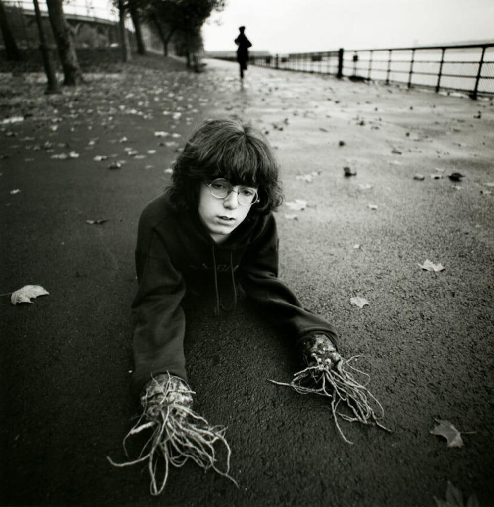 Black and white photo of a child with shaggy dark hair and glasses, lying on their stomach on a paved road strewn with leaves. Gnarled branches/roots emerge from their sweatshirt sleeves instead of hands