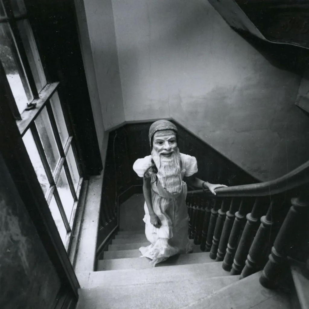 Black and white photo of a child in a light dress standing on a wooden stairway, shot from above. She wears a large mask of a man smiling menacingly with a long beard and cap.