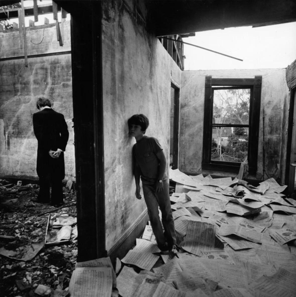 Black and white photo of a child in jeans and t-shirt leaning against a wall as if listening to the room behind it, in a broken-down building with fallen ceilings and floors strewn with newspapers and debris. Beyond the wall we see a man in a dark coat standing with his back to us, head bent, hands behind his back.