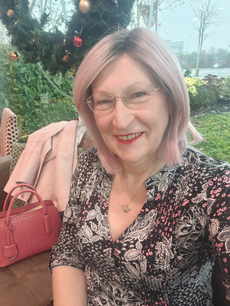 A pink haired woman sitting in a cafe wearing a black, white and pink leaf and small flower pattern v neck top, a gold bee pendant necklace and gold and crystal drop earrings.
A dark pink handbag and a light pink coat are beside me.