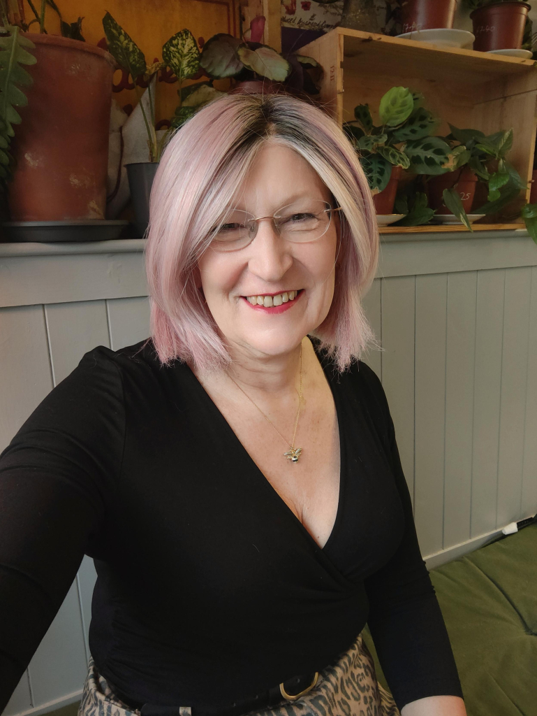 A pink haired woman sitting in a cafe wearing a black wrap top with an animal print denim skirt, a gold bee pendant necklace and gold hoop earrings.
There are plants on the shelf behind me.