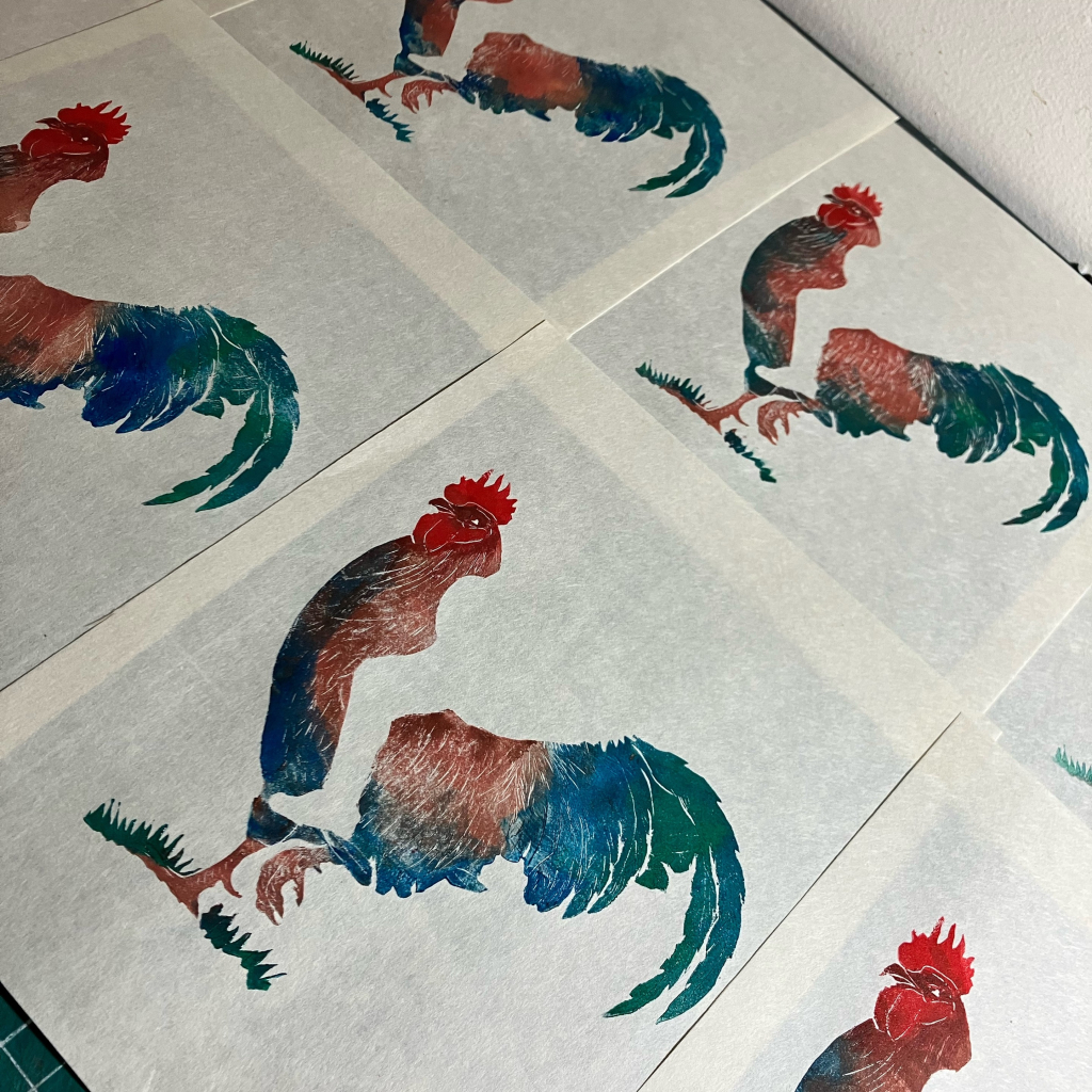 A collection of at least 6 Lino block prints on white 8” x 10” Japanese paper each with a rooster viewed from the side. The rooster has a red crest, brown body and feet with blue and green under- and tail feathers. There’s a little green grass by its feet. One foot is lifted like it’s walking forward. There’s an unprinted silhouette of a leg and boot, like a little man will later be riding the rooster like a horse. 