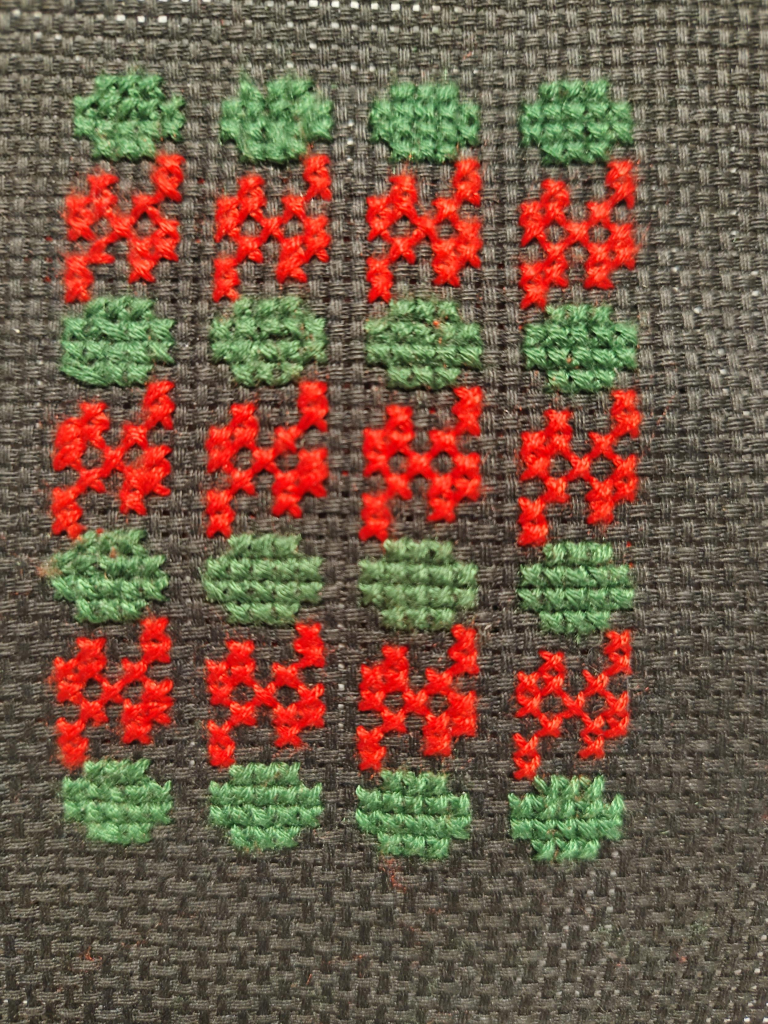 Close up of tatreez cross-stitch embroidery on coarse black fabric.

There are four columns, each with four green watermelons separated by red vines.

The quality of the stitching starts off rather poor but improves somewhat going from left to right.