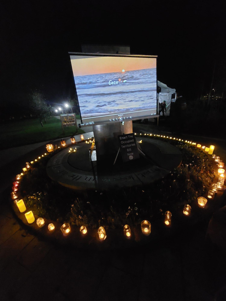 A screen with a film about Gaza projected on it outside Burnaby Park in Greystones at nighttime there are candles and tea lights in a circle on the floor