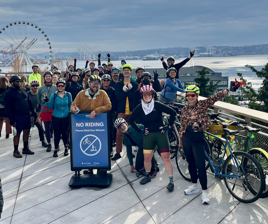 a group shot of a cycling club on the newly constructed concrete Seattle waterfront. There are about 25 of us willing to appear and we are all wearing our riding gear of many colors with bike helmets on. Several of those in the back are taking advantage and throwing out their arms to get attention. Your humble poster is simply smiling at the camera on the left with his hands behind his back