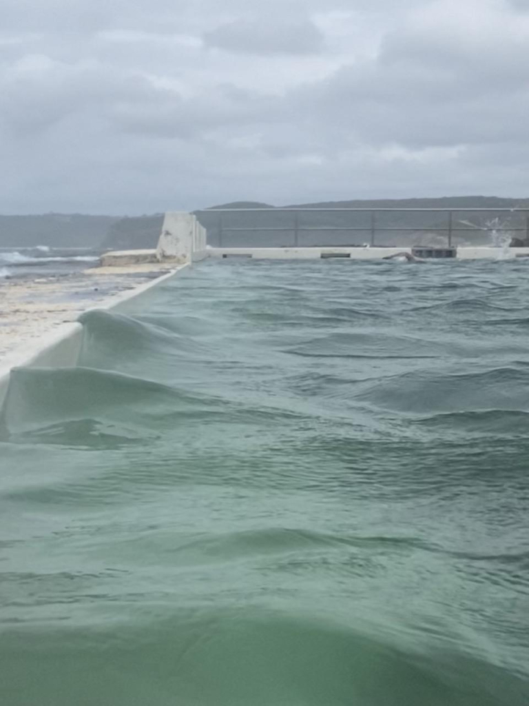 Photo looking southerly towards the back corner with the lap lane markers. The water near the white wall is almost an emerald green; small waves are reaching up to the edge of the wall. In the background, a swimmer is doing freestyle, one arm bent above the water and splashing from the direction of her feet. Clouds over the distant headland behind the baths. 