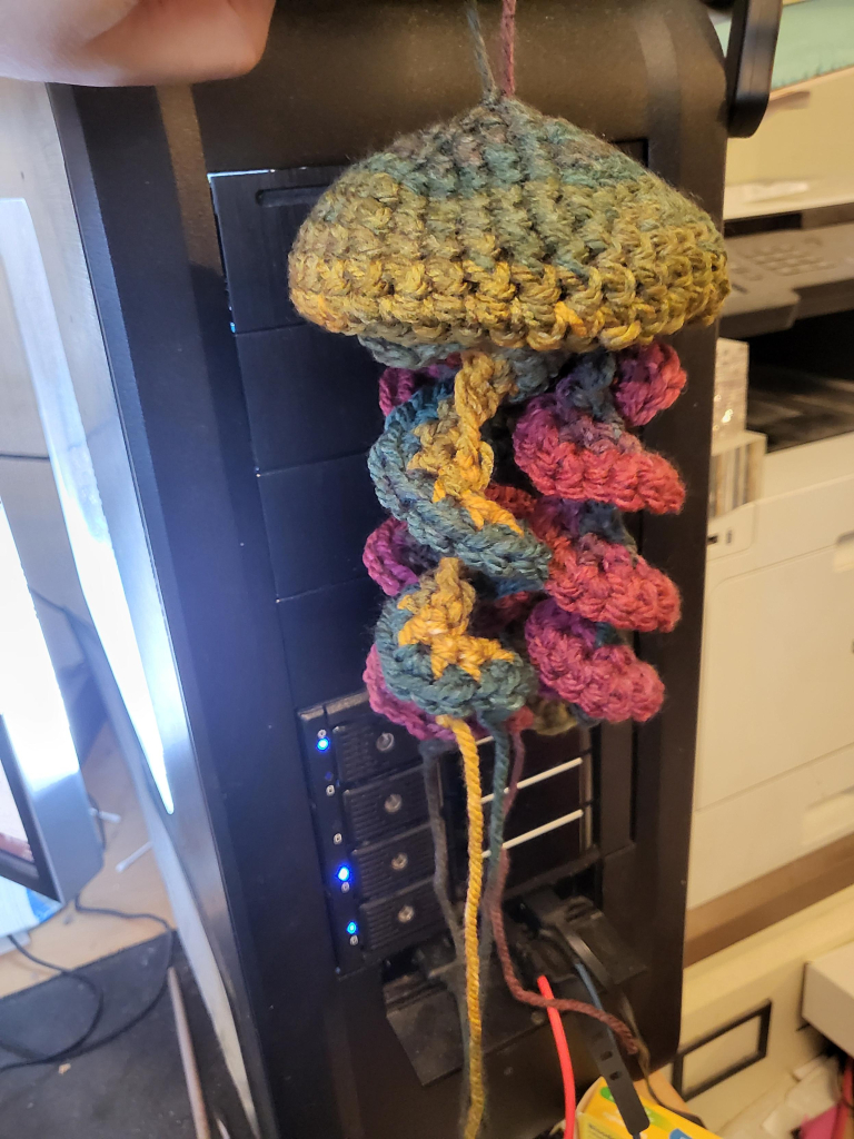 A multicoloured crocheted jellyfish, greenish body with yellow, blue, and pink tendrils. 