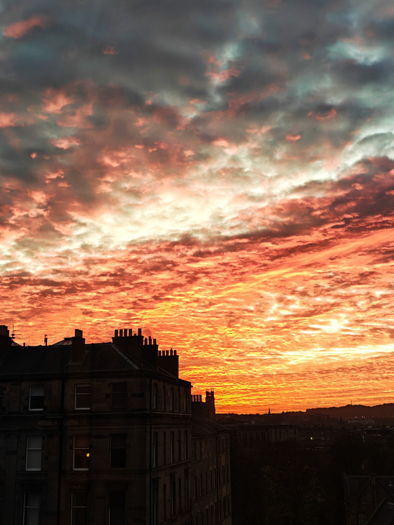 A photograph of the evening sky, with "mackerel" clouds tinged orange, pink and red. At the bottom left of the photograph is a silhouette Georgian tenement building.