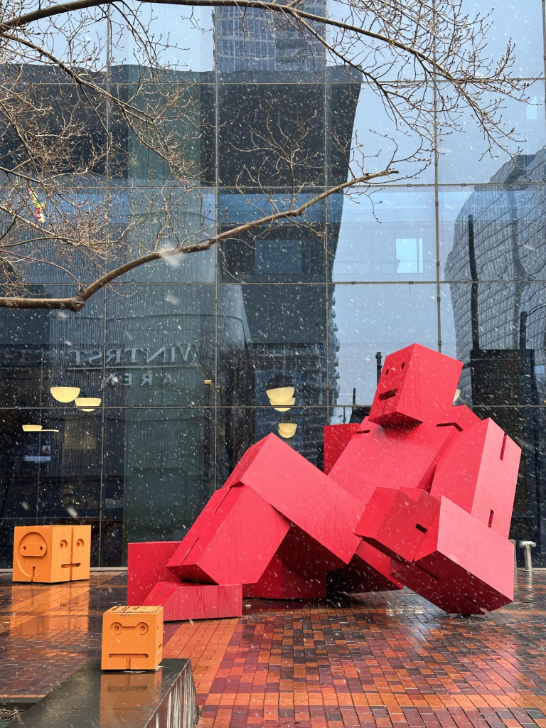 A huge robot sculpture composed of big red blocks sits in a plaza. Next to it are two yellow blocks with smiley faces on their sides. 