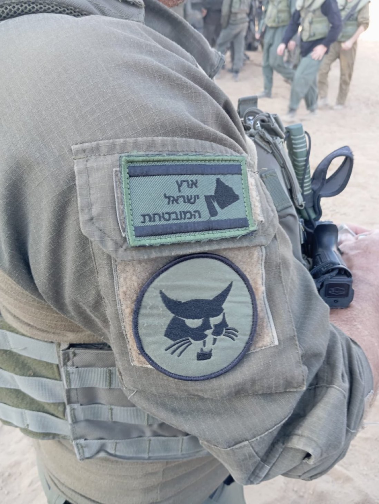 A soldier's patches, including one with a cat and one with Hebrew text and the shape of a country. More soldiers in the background.