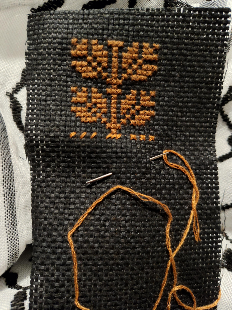 Close up photo of a strip of black aida cloth 11 count fabric that has two iterations of the Habbat Binn / Coffee Beans tatreez motif cross-stitched onto it in brown thread. The motif has a centre stem with leaves branching of left and right symmetrical to each other.

The cloth is propped against a white and black keffiyeh used as a background.