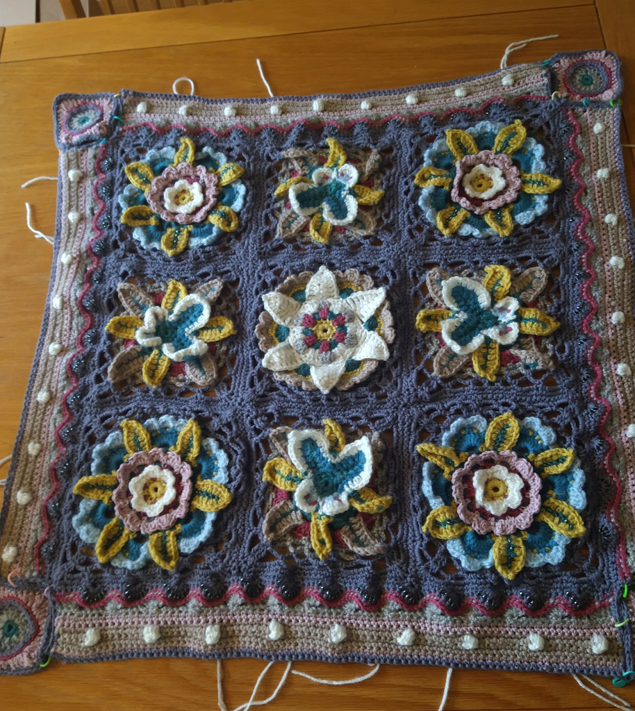 Nine crochet squares with grey background there are four corner flower designs in the centre of each row is a butterfly motif and the central square has a white flower design 