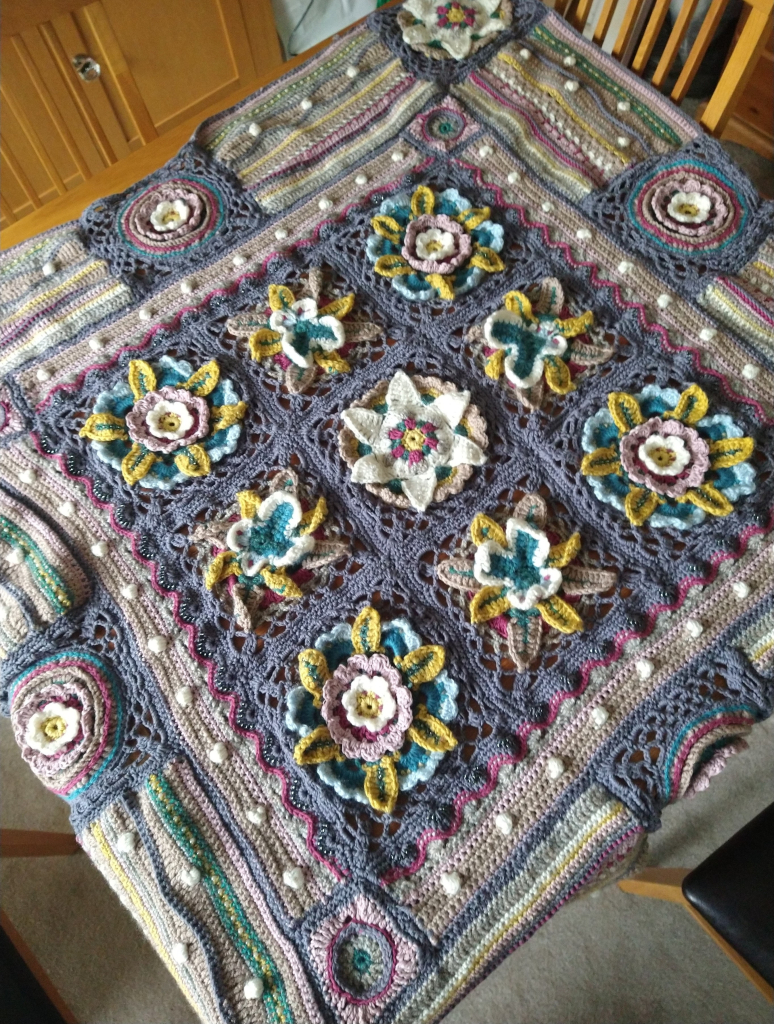 Crochet Afghan in shades of grey and pink viewed on an angle from above, the nine central squares has flowers or butterflies in the middle 