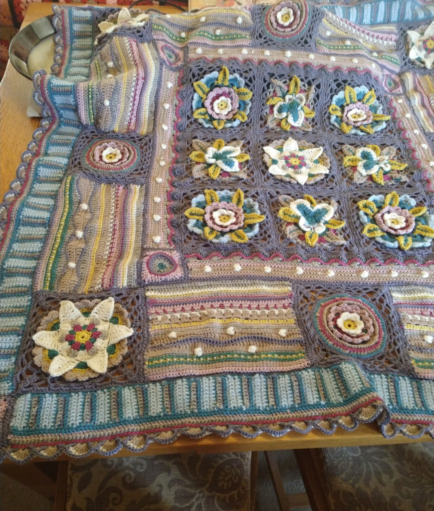 Jane Crowfoot  crochet Afghan, laid out on a table, it is in greys, blues, pinks, with flower and butterfly motifs 