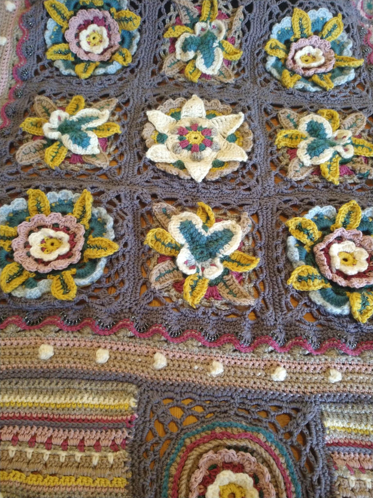 Close up of Jane Crowfoot  crochet Afghan, laid out on a table, it is in greys, blues, pinks, with flower and butterfly motifs, with bobble and bead stitches 