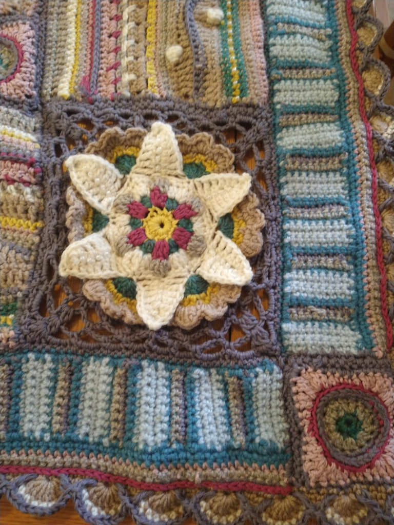 Close up of Jane Crowfoot  crochet Afghan, laid out on a table, it is in greys, blues, pinks, with flower motif,