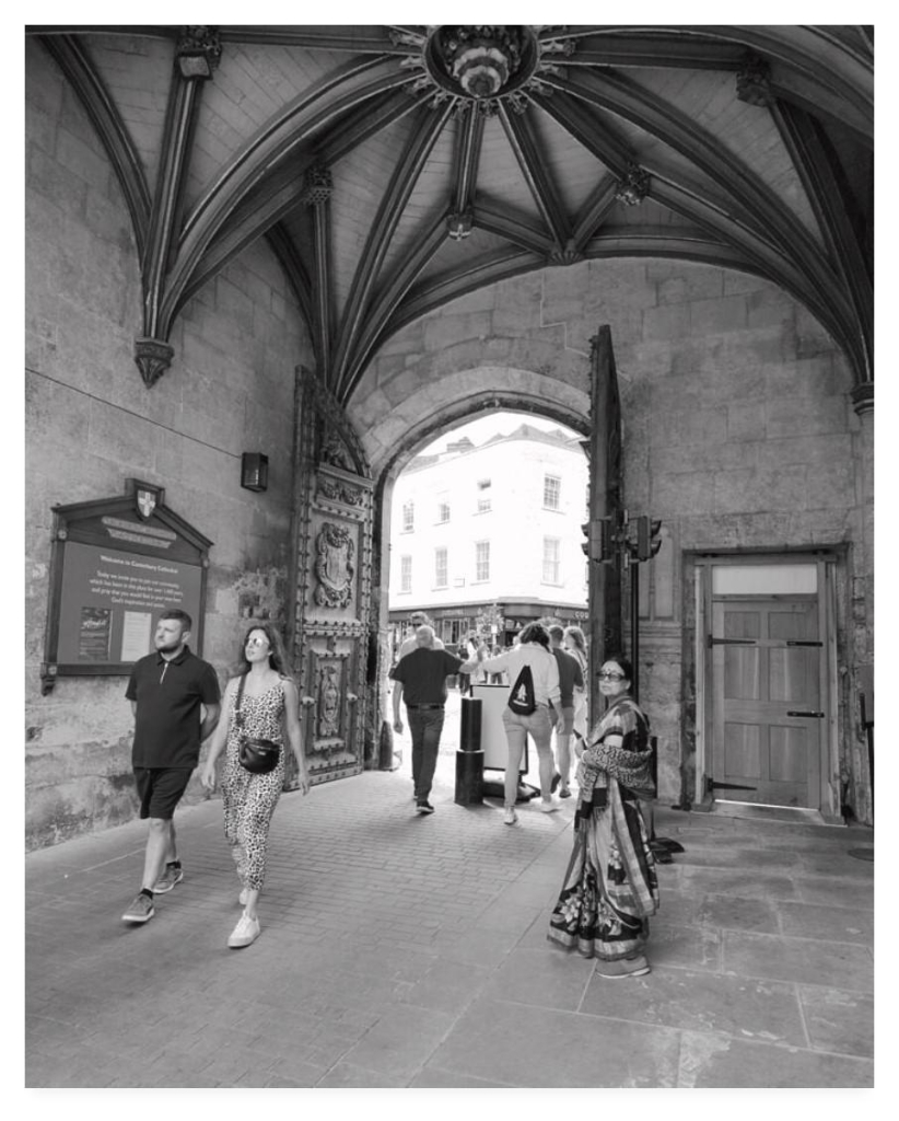 Black and white portrait photograph showing the inside of the Christchurch Gateway at Canterbury Cathedral.  The ornately carved wooden doors are open allowing people to roam through.