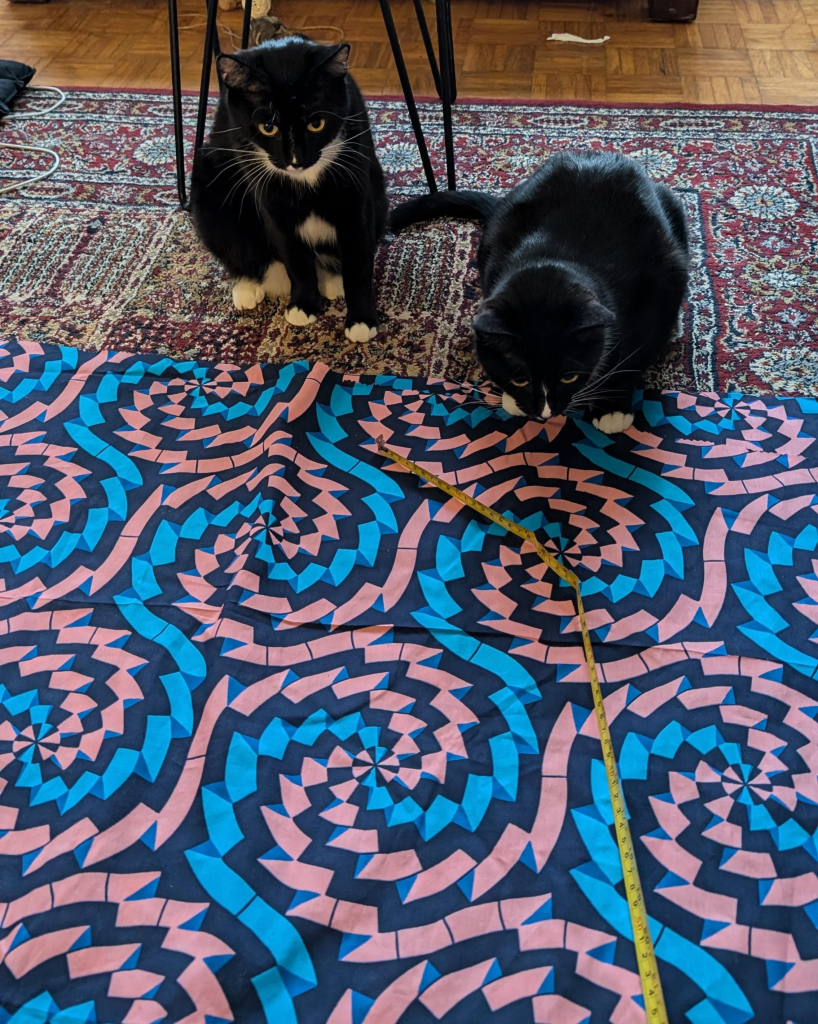 Photo of two black and white kittys looking down at a huge piece of pink, black and blue fabric they are sitting close to. They're also looking at a tape measure which has been pulled out and laid on the fabric 