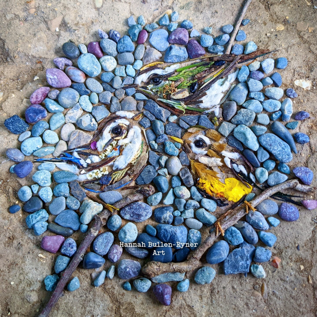 A piece of street art made from feathers, coloured stones and sticks.  It shows three birds - two warblers and a golden-crowned kinglet, coloured with feathers.  Each is perching on a stick, against a circle made of tiny pebbles in numerous shades of blue and purple.