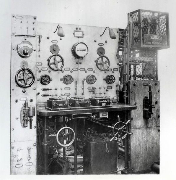 An antique control panel, with numerous gauges, hand cranks, and large switches. To the right is some component in a cage, with a warning regarding high voltages.