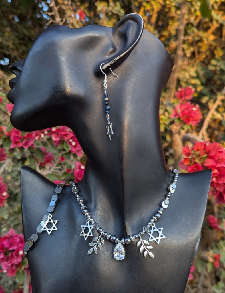 A handmade set of Magen David necklace bracelet and earrings 
Earrings with sapphire gemstones and sterling silver hooks and Magen David stainless steel pendants 
Necklace with silver leaves and zirconia drop and stainless steel Magen David, hematite styled beads and acrylic shining beads
Bracelet with hematite styled beads and stainless steel Magen David 

On a black doll, in the background are pink bush flowers