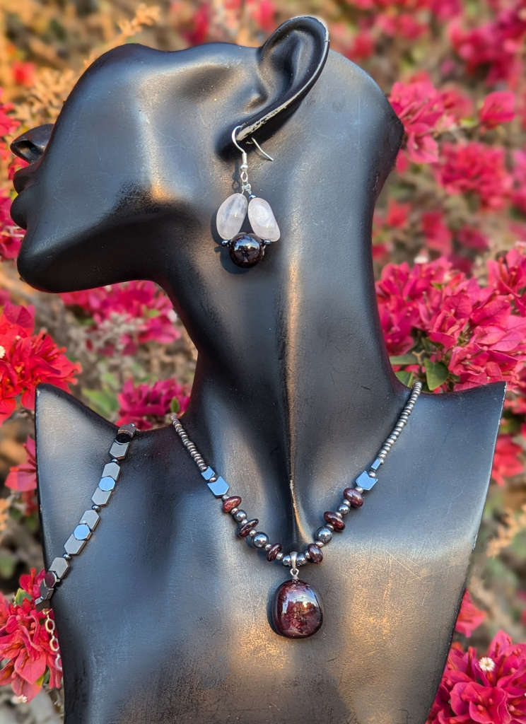 On a black doll with pink flowers bush background 
handmade Garnet set, featuring a unisex necklace and bracelet with Garnet gemstones and Hematite styled beads, and Garnet and Rose Quartz gemstones earrings on sterling silver hooks