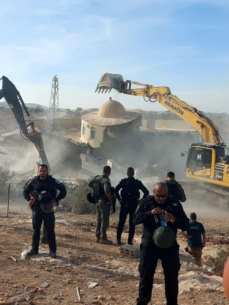 Police officers standing and watching a large bulldozer destroying a mosque. 