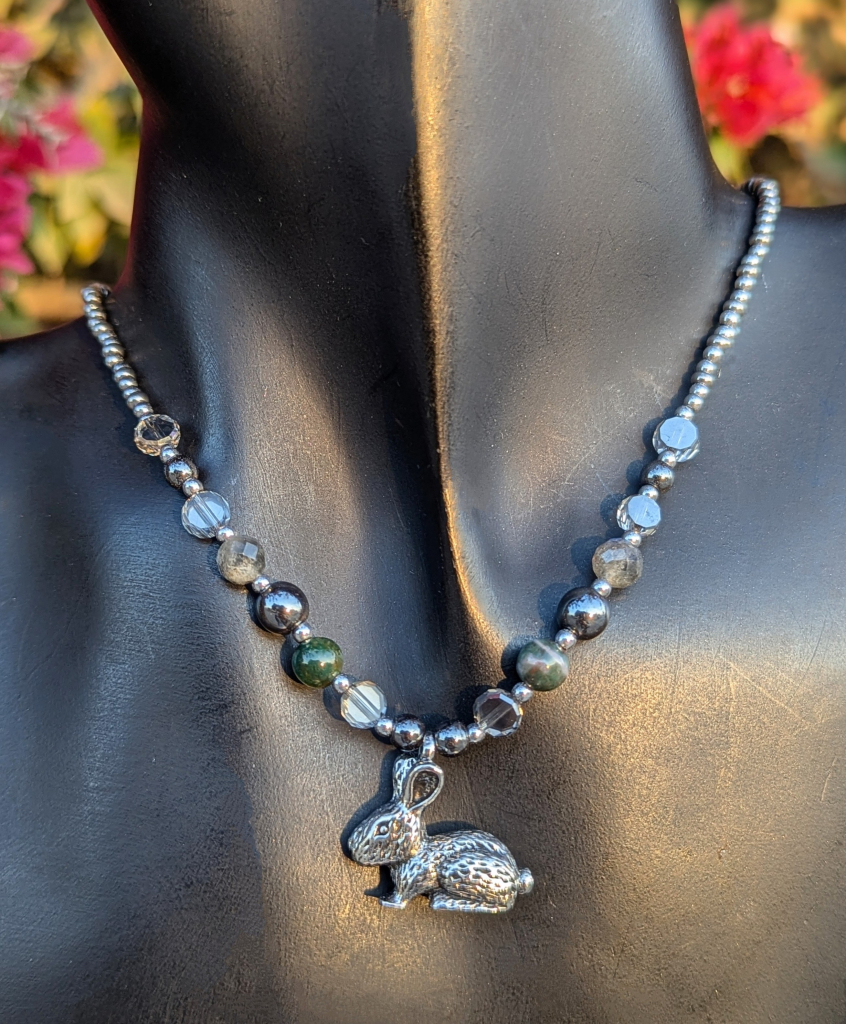 Handmade necklace with green agate gemstones, labradorite gemstoned, hematite styled beads and grey see through acrylic beads and silvery metallic beads with a bunny pendant 
On a black doll
Pink flowers bush in the background 
Close up on necklace