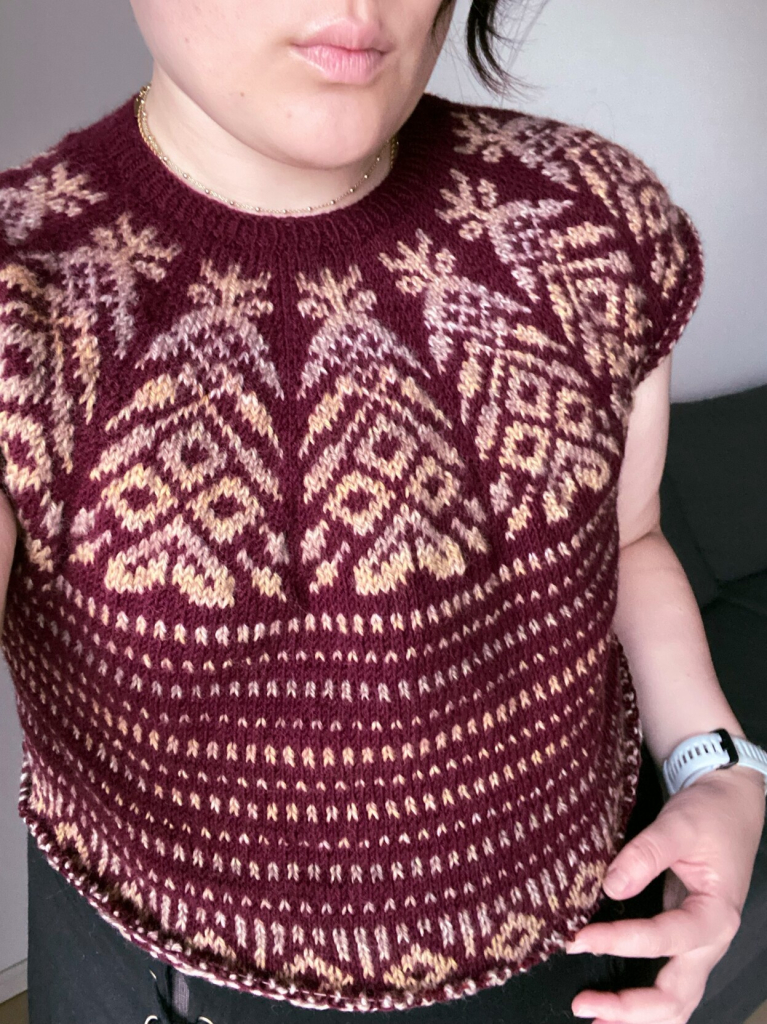 A close-up of a person wearing a maroon knitted sweater with intricate colorwork patterns in shades of beige and light yellow. The sweater features a rounded neckline and short sleeves. The person's face is partially visible.