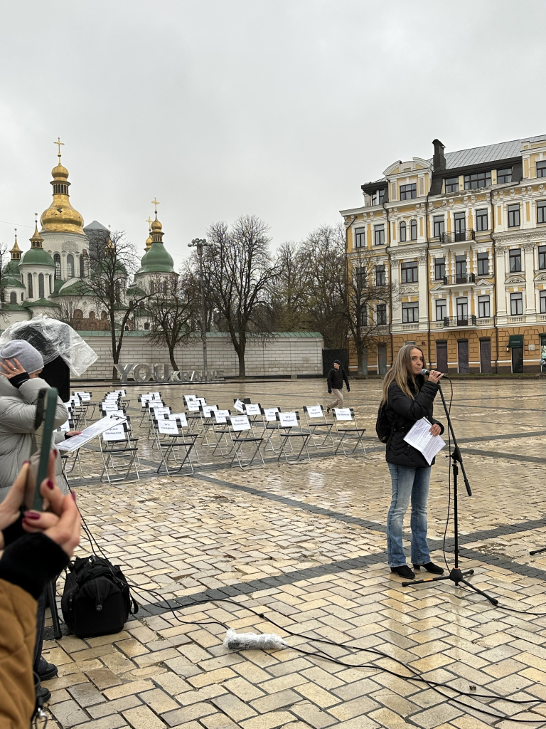 A view of the empty chairs on Sofiivska Square. In the background is the cathedral. In the foreground, the wife of a prisoner speaks  it’s a gray, drizzly day. 